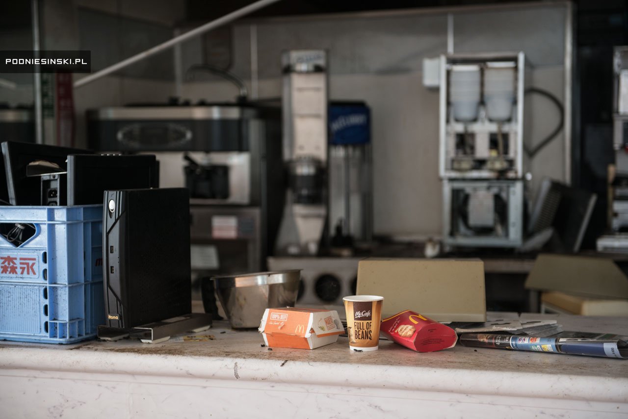 Mostrador de un McDonald's abandonado: entre el polvo quedan una caja de hamburguesa, un vaso de McCafé y una bolsa de patatas, con las máquinas de café apagadas al fondo.