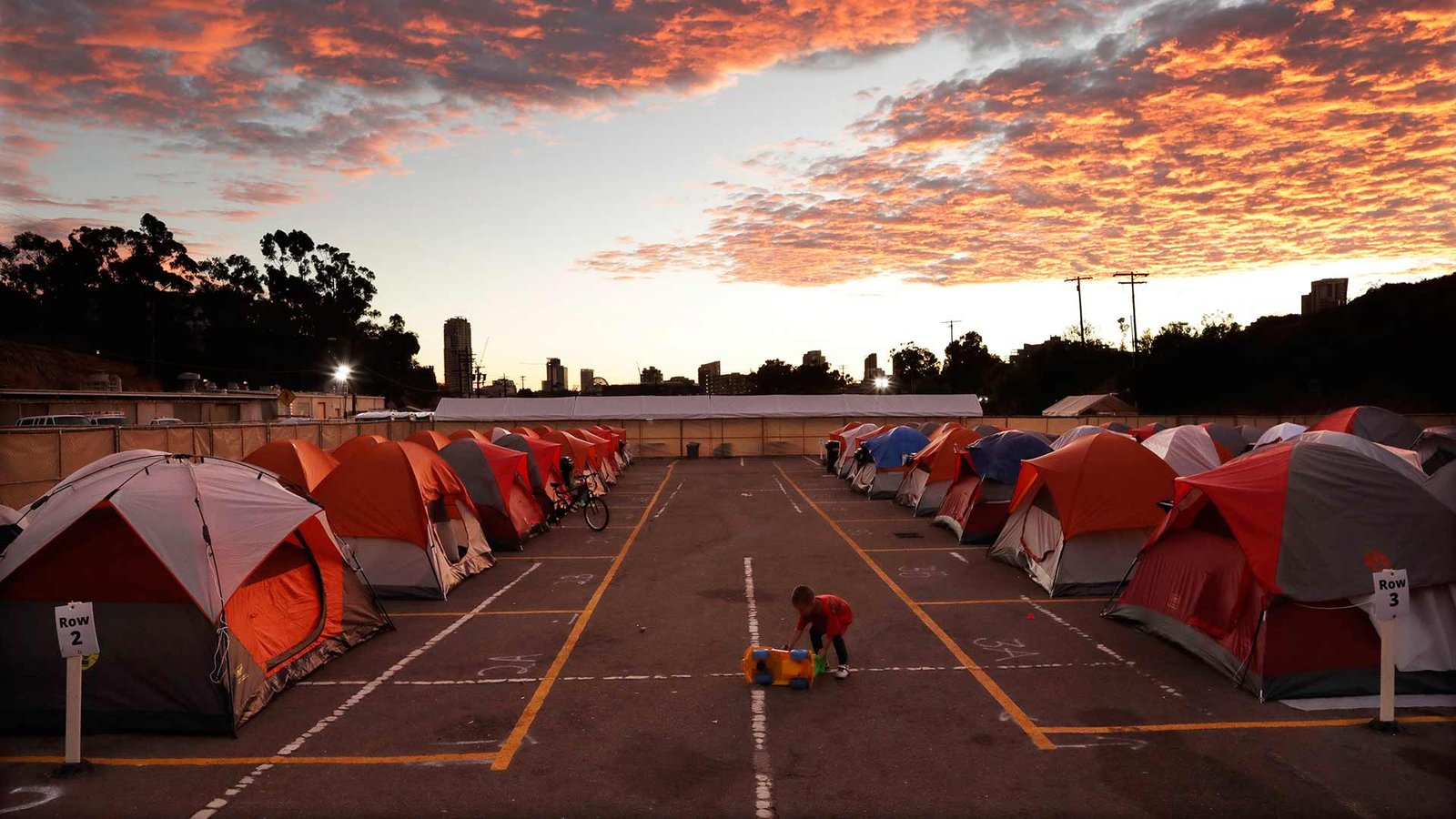 Aparcamiento al aire libre en San Diego convertido en campamento: dos hileras de tiendas de campaña ocupan las plazas marcadas en el asfalto, señalizadas con carteles de «Row 2» y «Row 3», mientras un niño empuja un camión de juguete por el pasillo central al atardecer, con el perfil de la ciudad al fondo.