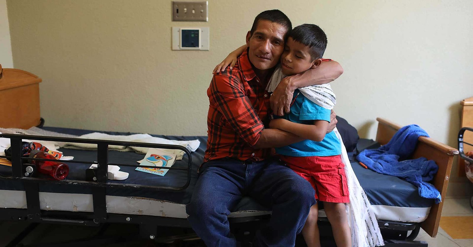 Un hombre con camisa de cuadros abraza a un niño sentado en su regazo, en una habitación con dos camas metálicas y un cuaderno de colorear sobre la sábana.
